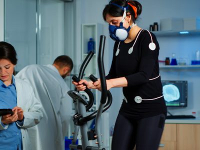 Woman athlete with mask running on cross trainer in sports science lab measuring performance and oxygen consumption during Vo2max test. Medical researcher looking at EKG scan monitoring heart rate